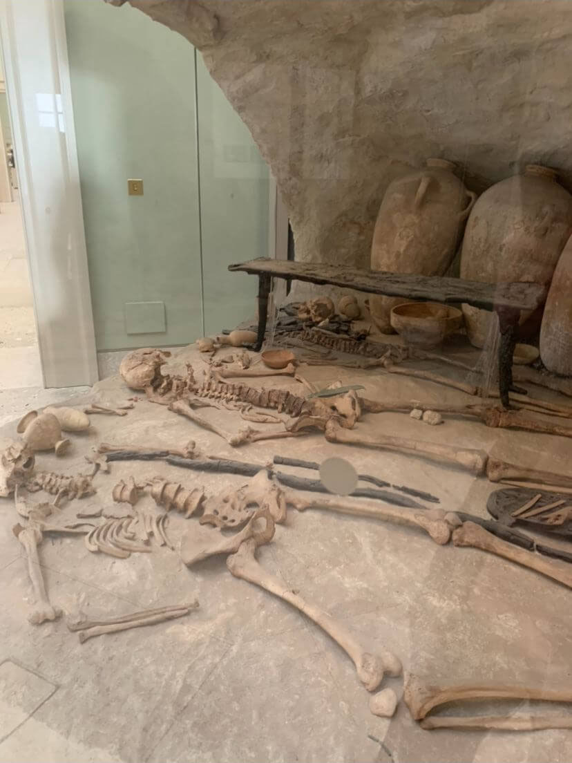 Two skeletons lay on a sandstone ground. There is a wooden bench to the right of two skeletons, and a third skeleton after the bench. To the right of all three skeletons are big ceramic pots and a ceramic bowl. They are all kept behind a glass wall.